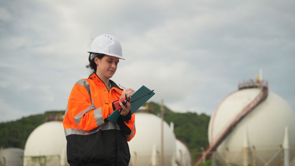 A female utility worker wearing a white hard hat and high-visibility orange jacket, holding a clipboard and radio while inspecting industrial storage tanks in an outdoor facility.